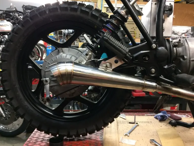 Mechanic comparing two exhaust pipe ends on a workbench, with clamps and a tape measure.